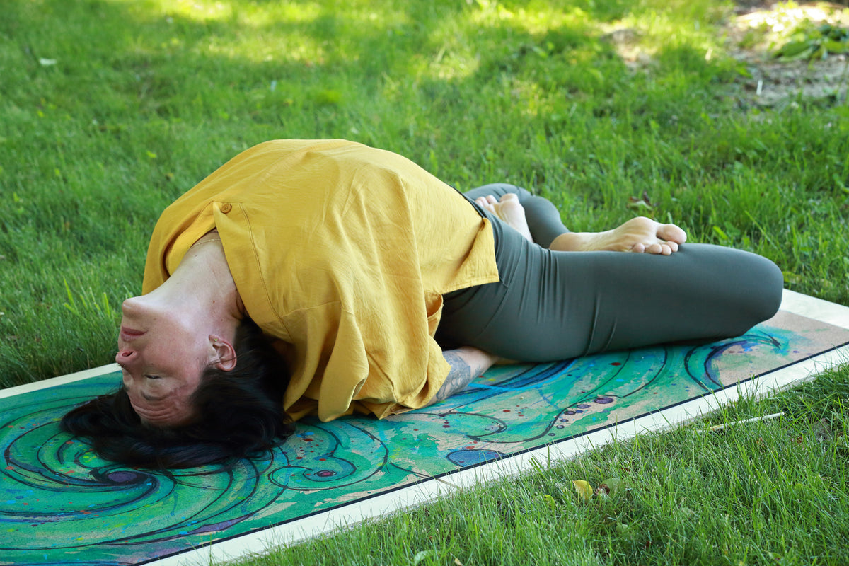 A woman doing yoga outside, practicing  matsyasana, fish pose, outdoors on a grassy lawn, lying on a colorful yoga mat in a deep forward bend pose. The person is wearing a mustard yellow top and dark gray leggings.