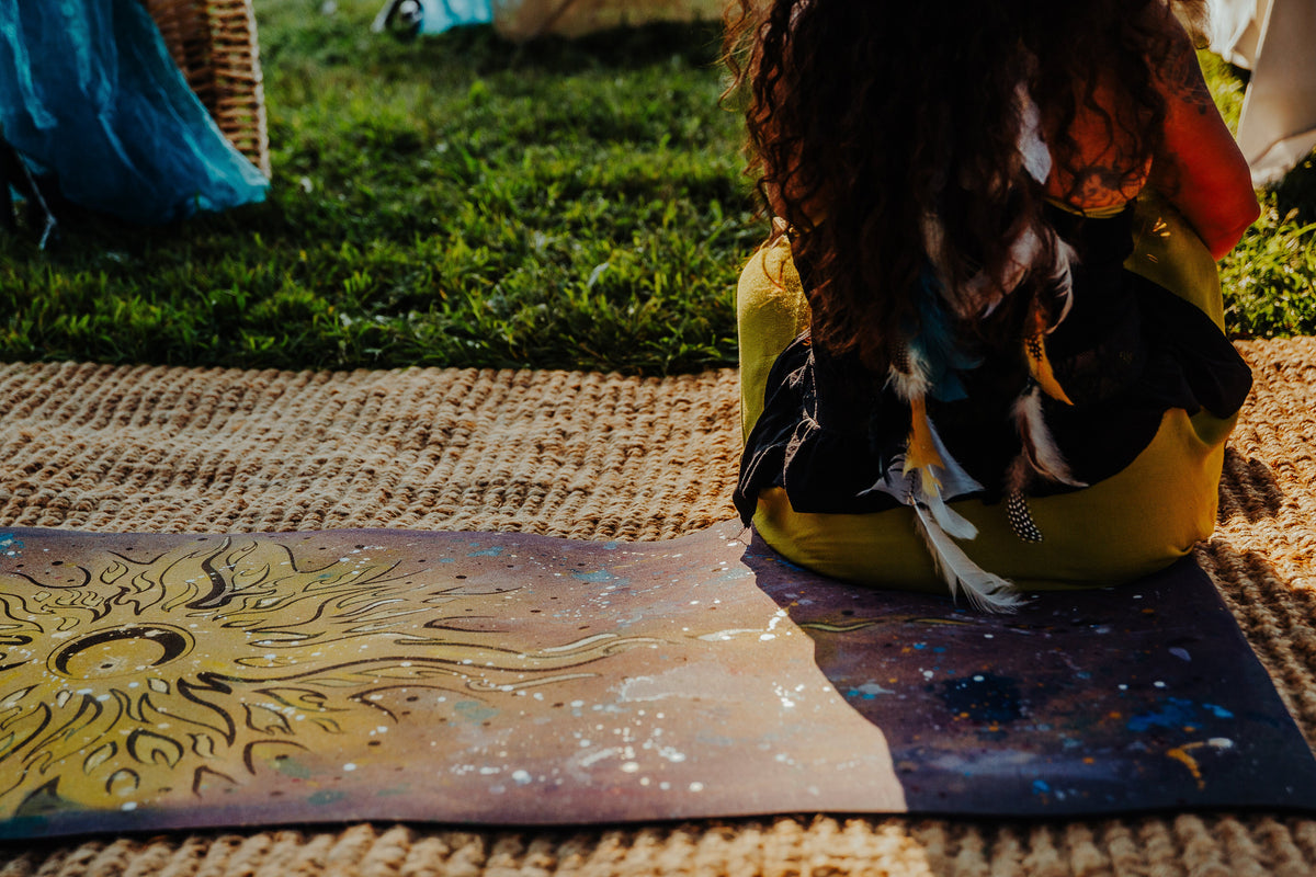 A person sits outdoors on a woven juite rug on a colorful hand painted cork yoga mat featuring a stylized sun design, with grass and other outdoor elements in the background.